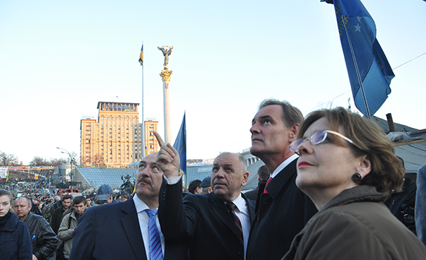 Several people look upwards; buildings and flags in the background.