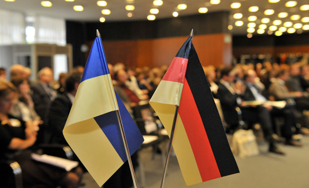 Eine deutsche und eine ukrainische Tischflagge im Vordergrund; unscharf dahinter eine Gruppe von Menschen in einem Saal.