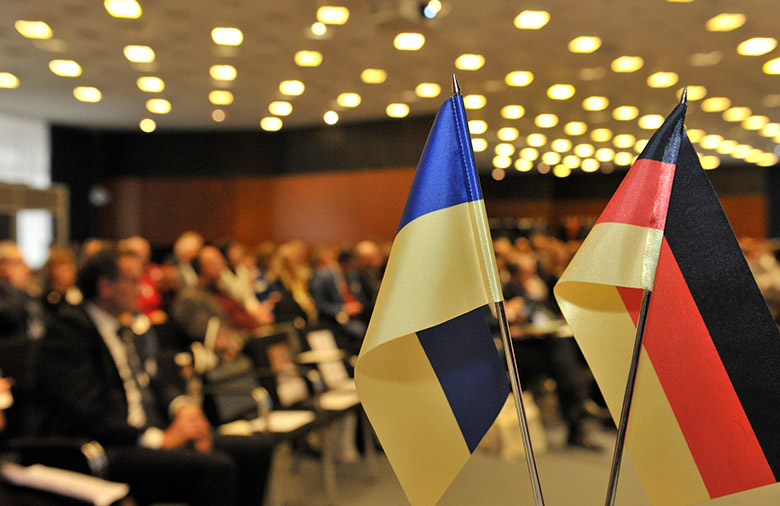 Im Vordergrund eine deutsche und eine ukrainische Flagge; im Hintergrund Personen in einem Saal.