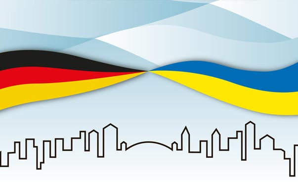 Eine gemalte deutsche und eine ukrainische Flagge schweben über einer Stadtsilhouette.