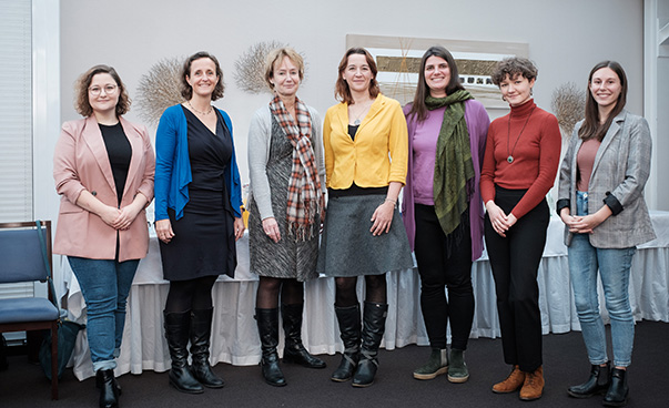 Sieben Frauen stehen nebeneinander in einem Raum und posieren für die Kamera.