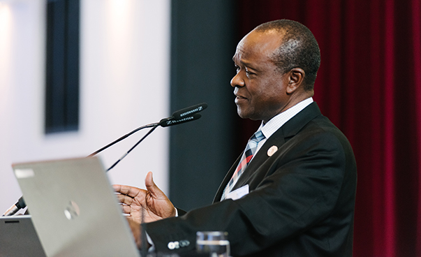 TeilErastus J. O. Mwencha ist von der linken Seite aus zu sehen, wie er vor einem Rednerpult mit Mikro steht und spricht. Dabei gestikuliert er mit seiner rechten Hand.nehmende tauschen sich während der Konferenz aus.