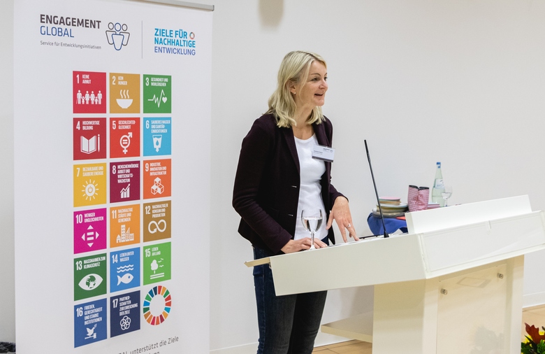 Eine Frau steht am Rednerpult. Links neben ihr ein Poster mit den Symbolen der 17 Nachhaltigkeitsziele.