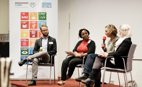 Drei Frauen und ein Mann sitzen auf dem Podium; im Hintergrund ist eine Stellwand mit den Logos der globalen Nachhaltigkeitsziele zu sehen. Eine der Frauen spricht in ein Mikrophon.