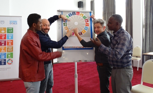 Vier Männer posieren vor einem Flipchart mit den Symbolen der globalen Nachhaltigkeitsziele.