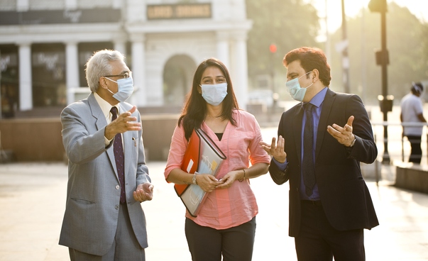 Zwei Männer und eine Frau mit Mund-Nase-Schutz gehen diskutierend über einen städtischen Platz.