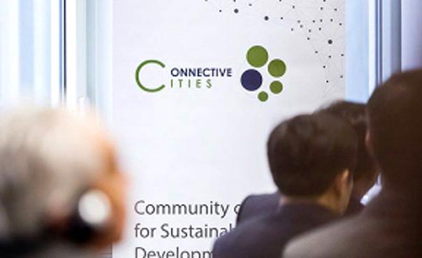 Auf einem großen Banner auf einer Bühne ist das Connective Cities Logo zu sehen. Im Vordergrund sind umschafft die Hinterköpfe mehrerer Personen zu sehen.