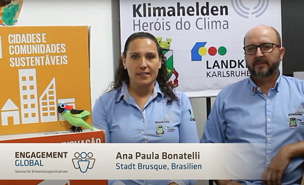 Ana Paula Bonatelli von der brasilianischen Stadt Brusque spricht in die Kamera, neben ihr steht ein Mann. Im Hintergrund sind verschiedene Palkate zu sehen.