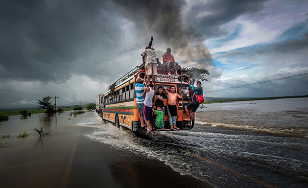 A bus, with several people clinging to the outside of the back, drives over a flooded road; very dark clouds can be seen in the sky.