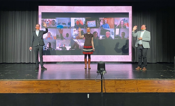 There are three people on a stage. In the background there is a large screen with other people logged in.