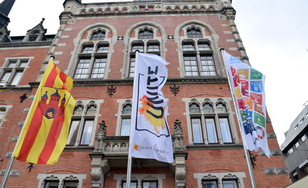 Vor einem historischen Gebäude sind drei Flaggen an einem Fahnenmast zu sehen.