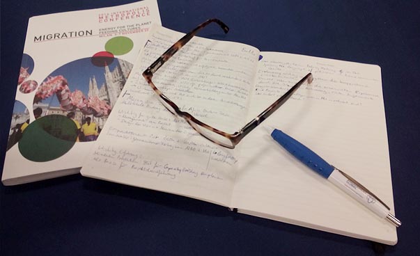 Auf eine Notizbuch liegen eine Brille und ein Kugelschreiber. Daneben eine Broschüre mit dem Titel Migration.