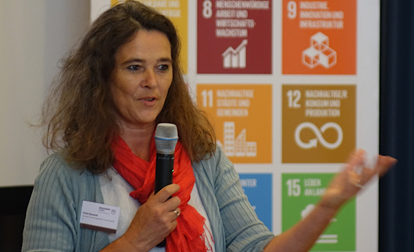 Eine Frau spricht in ein Mikrophon; im Hintergrund sind auf einer Stellwand unscharf einige Symbole der globalen Nachhaltigkeitsziele zu erkennen.
