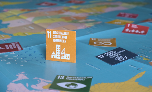 Aufgestelltes gelbes Icon von SDG 11 (Nachhaltige Städte und Gemeinden) auf einer ausgelegten Weltkarte. Ringsum liegen weitere SDG-Icons.