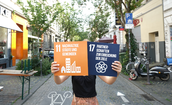 Eine Person steht in einer kleinen Kopfsteinpflasterstraße und hält Papp-Icons der SDGs 11 und 17 hoch. Die Schilder verdecken den Kopf.