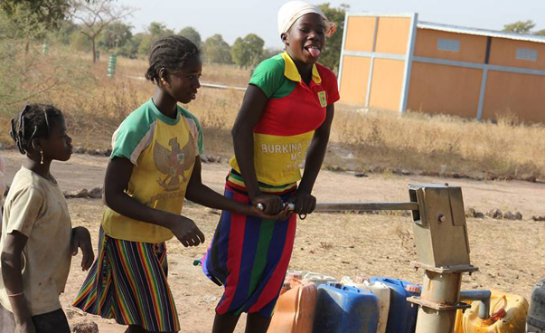 Drei Mädchen sind zu sehen; eine pumpt Wasser aus einer Pumpe; einige Kanister stehen daneben.