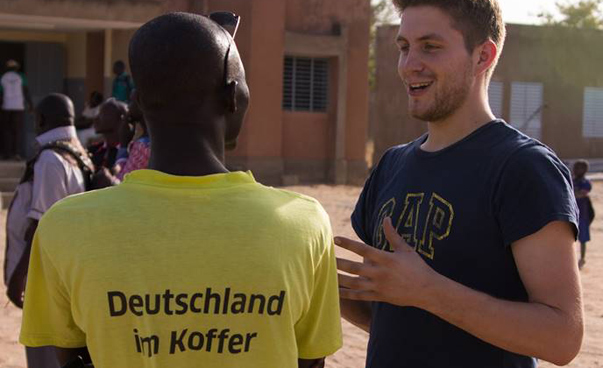 Zwei Männer unterhalten sich, der eine trägt ein T-Shirt mit der Aufschrift Deutschland im Koffer.