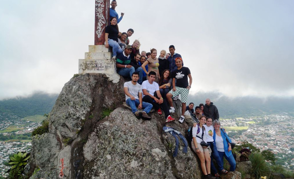 Viele junge Leute posieren um ein Gipfelkreuz herum; im Hintergrund ist eine Stadt zu sehen.