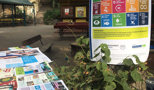 Auf einem Außengelände mit Bänken liegen Broschüren auf einem Tisch; rechts ist ein Plakat mit den globalen Nachhaltigkeitssymbolen zu sehen.