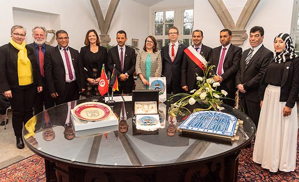 Elf Personen stehen um eine runden Tisch herum, auf dem unter anderem kleine Flaggen zu sehen sind, und lächeln in die Kamera.