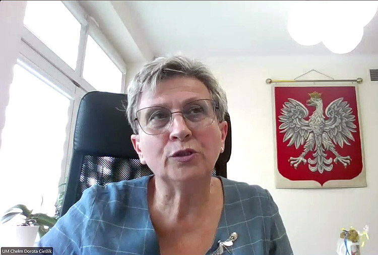 Dorota Cieślik, stellvertretende Bürgermeisterin der polnischen Stadt Chelm, per Video aus ihrem Büro hinzugeschaltet. Im Hintergrund das Stadtwappen von Chelm.